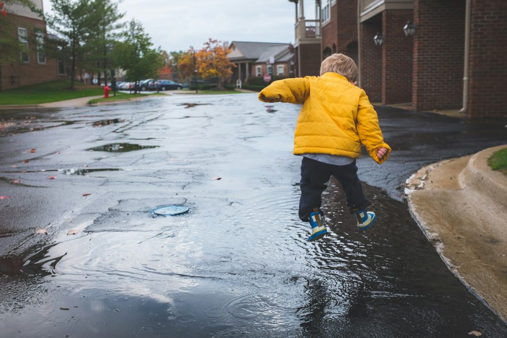 Enfant en doudoune jaune et bottes de pluie sautant dans une flaque un jour d’automne